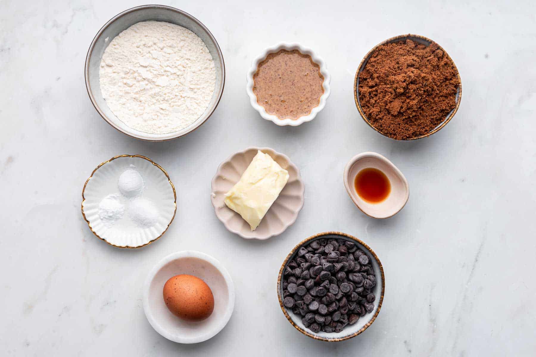 Bowl of flour, brown sugar, chocolate chips, egg and vanilla on white counter.