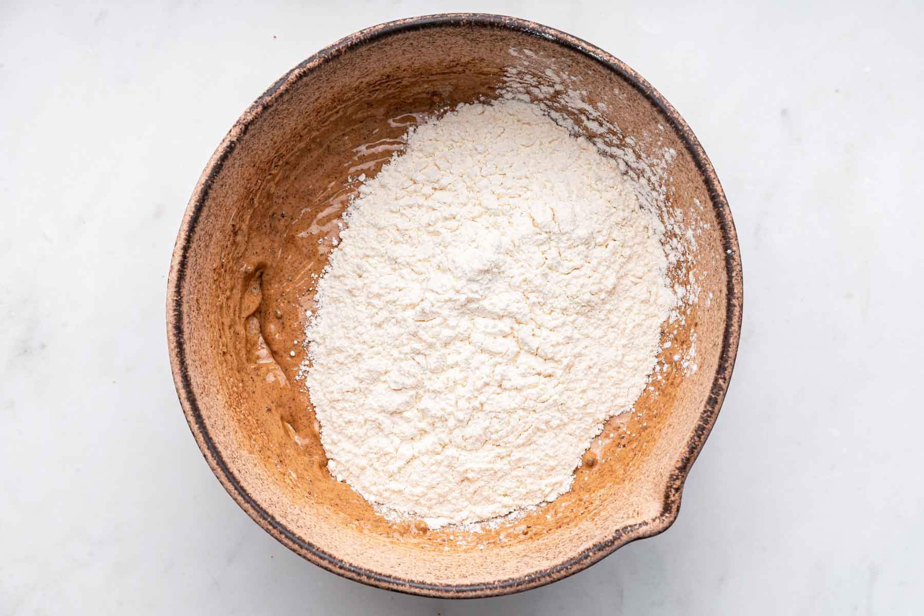 Flour and dry ingredients on top of brown dough in brown bowl.