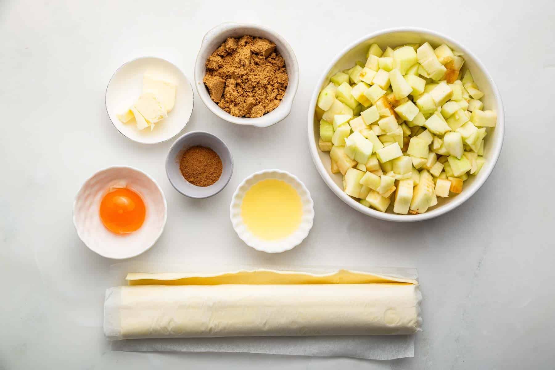 Bowls on counter with egg yolk, roll of dough, chopped fruit, and brown sugar.