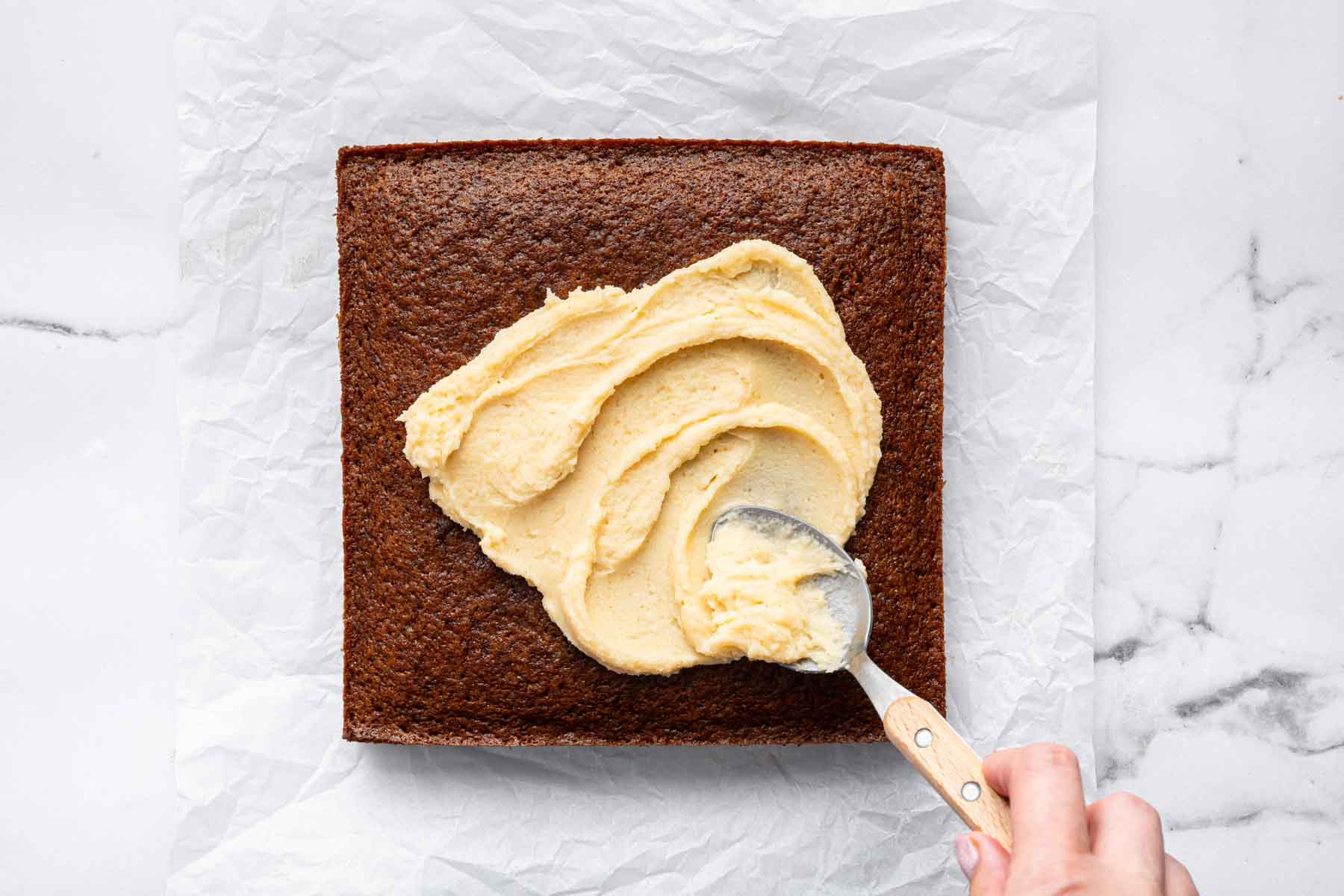 Square brown applesauce cake with caramel colored frosting being spread on top with a spoon.