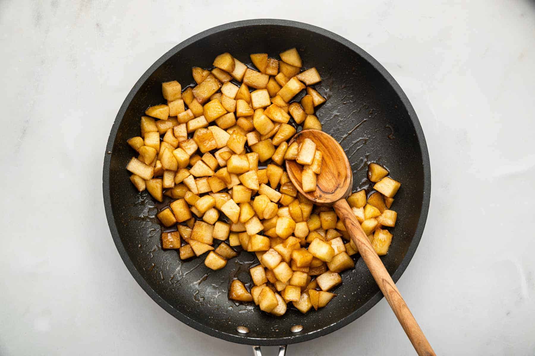 Black skillet with diced apples cooking and a wooden spoon.