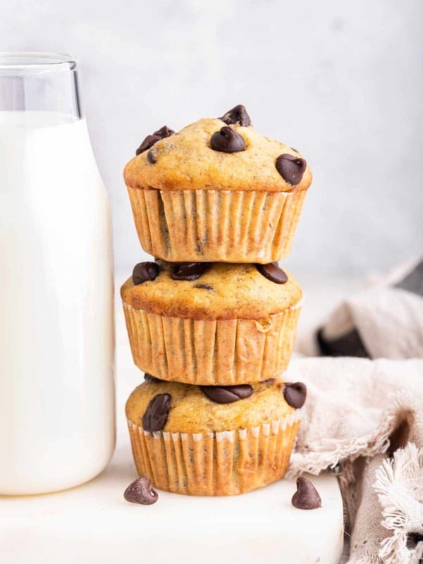 Three chocolate chip banana muffins stacked next to a glass of milk.