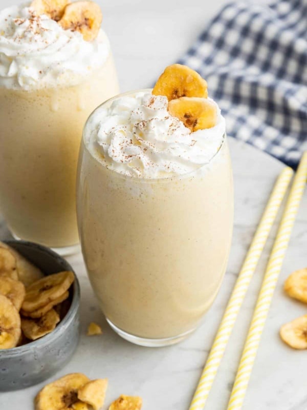 Two glasses of banana milkshake garnished with whipped cream and dried banana slices.