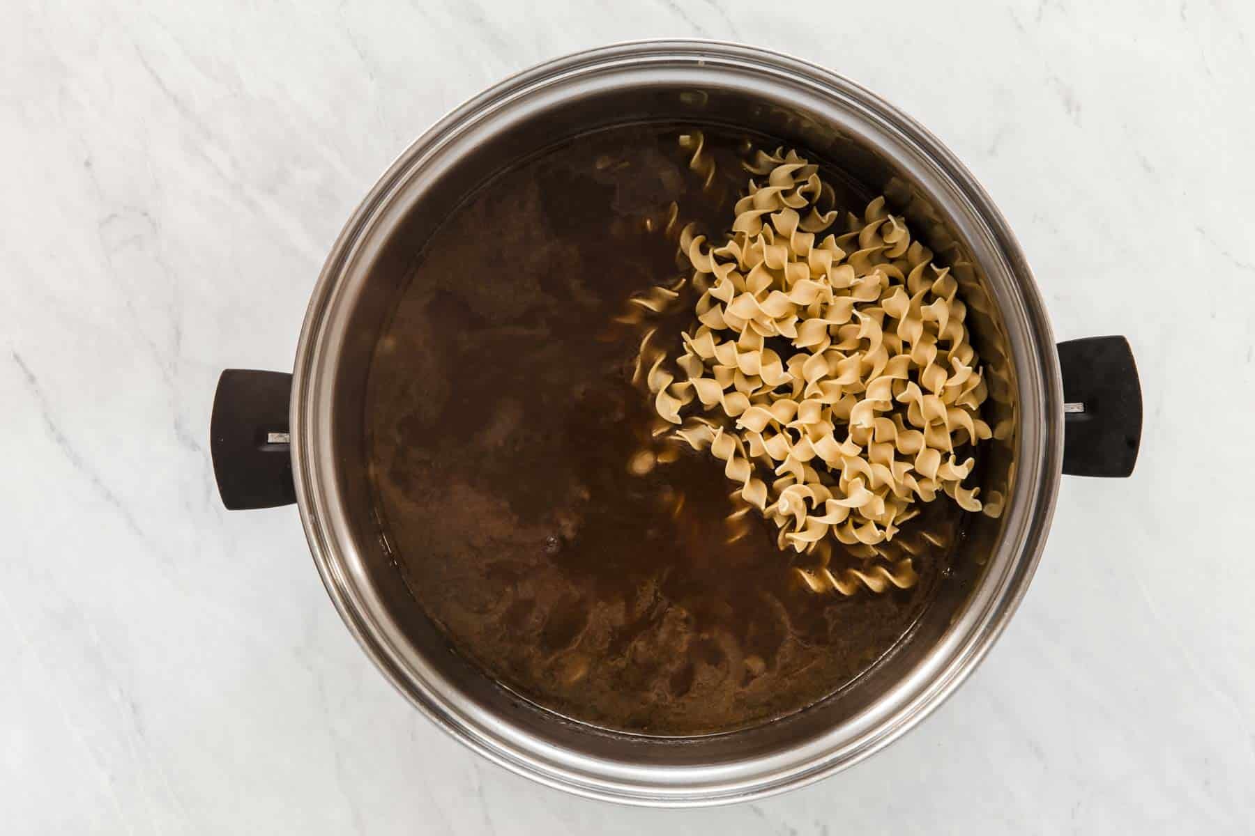 Round silver soup pot with brown liquid and egg noodles inside.