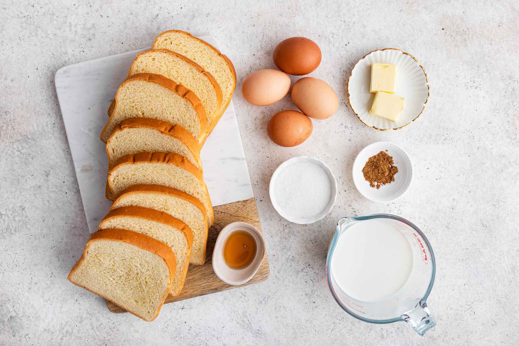 Sliced bread, eggs, syrup, cinnamon and milk on grey counter.