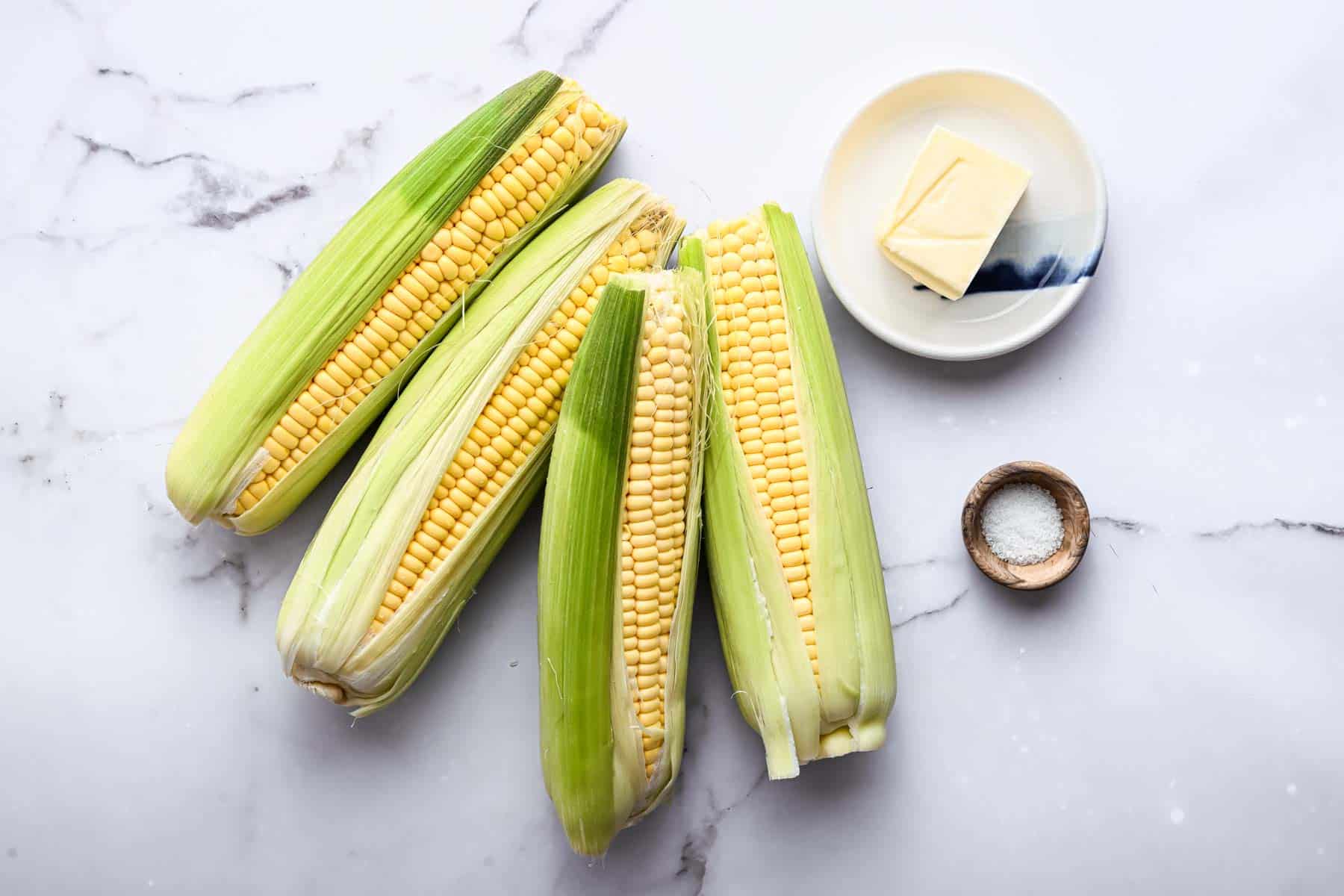 Four ears of corn with a plate of butter and salt next to it.