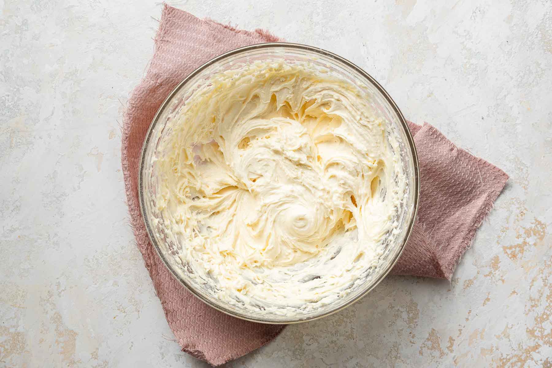 Whipped cream cheese frosting in clear bowl on pink towel.