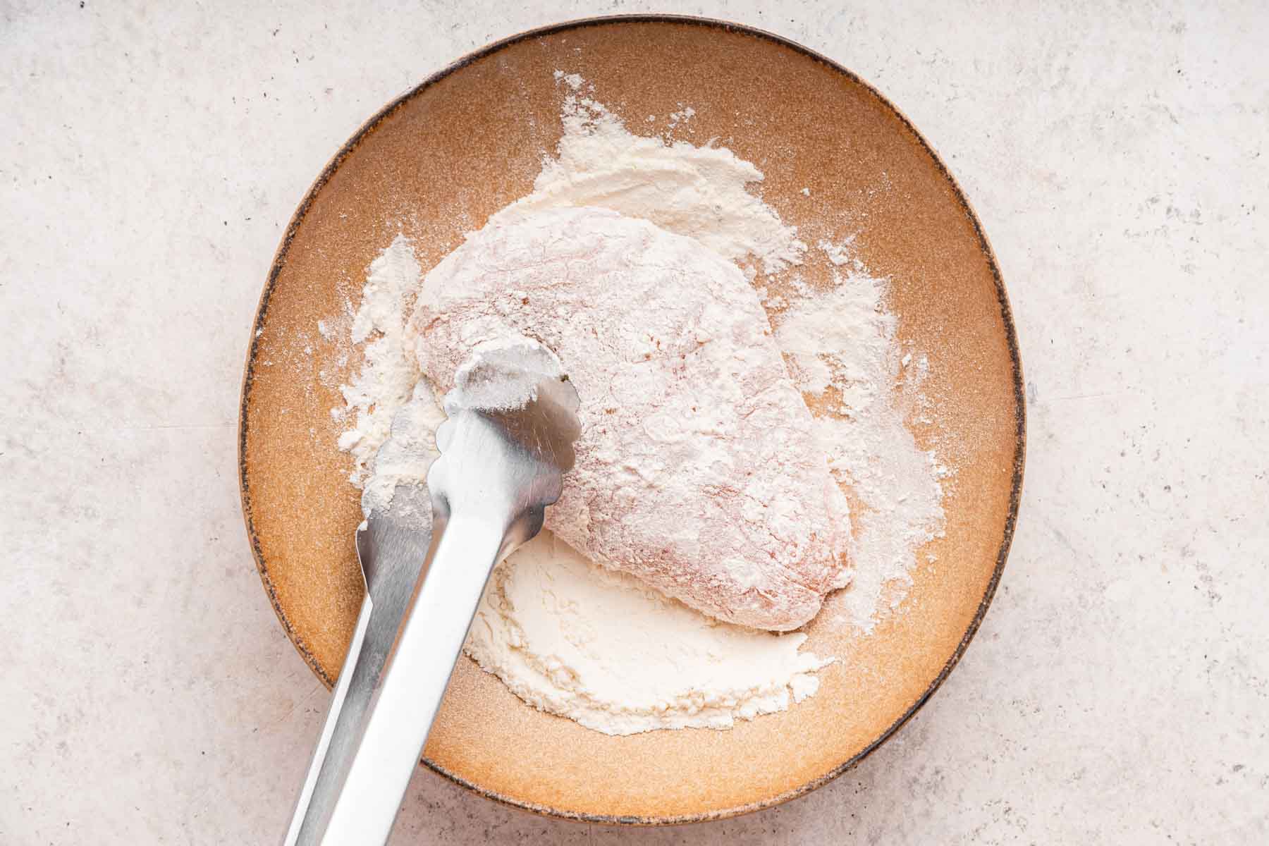 Tongs holding chicken being coated in flour.