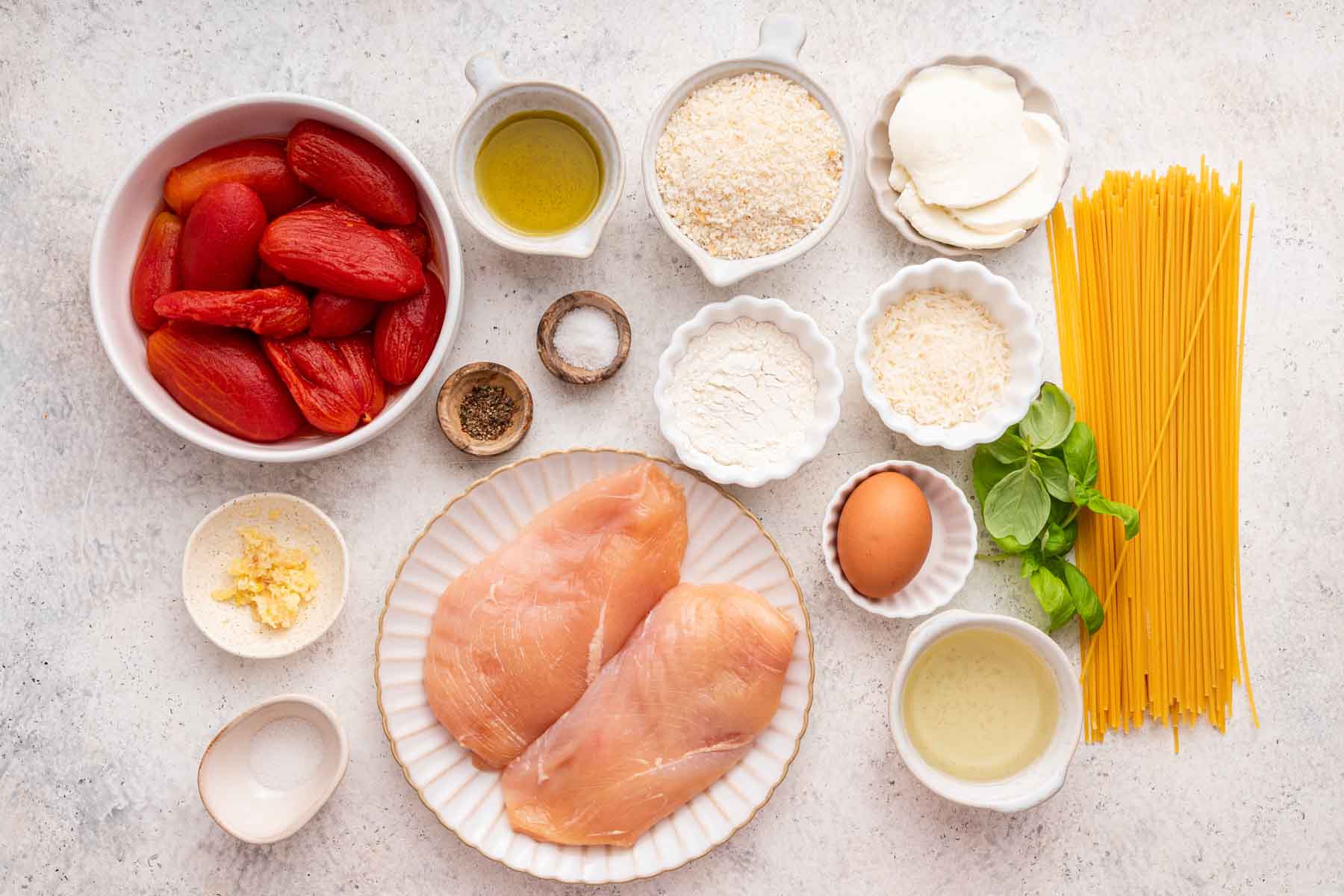 Bowls of canned tomatoes, chicken breast, noodles, and spices on grey counter.