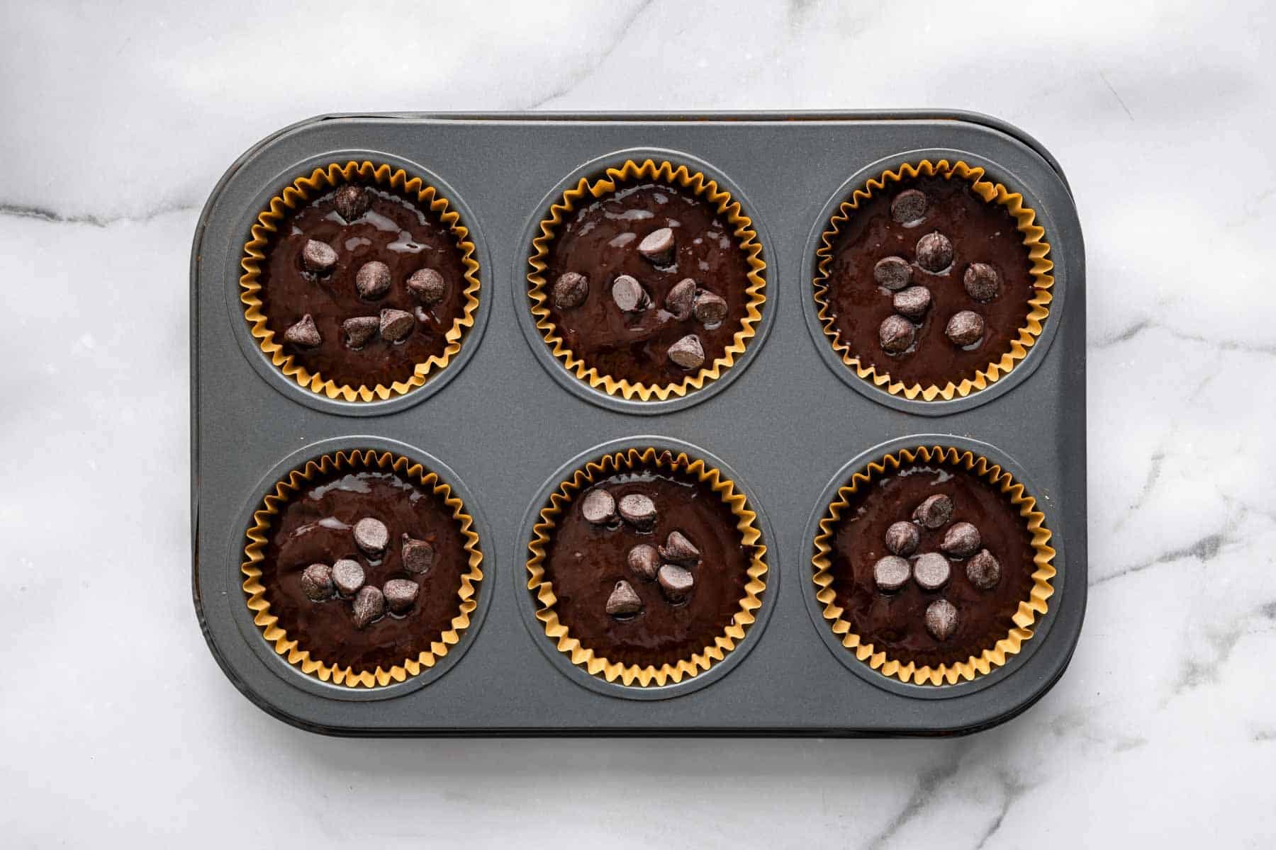 Raw chocolate muffin batter in muffin pan with chocolate chips on top.