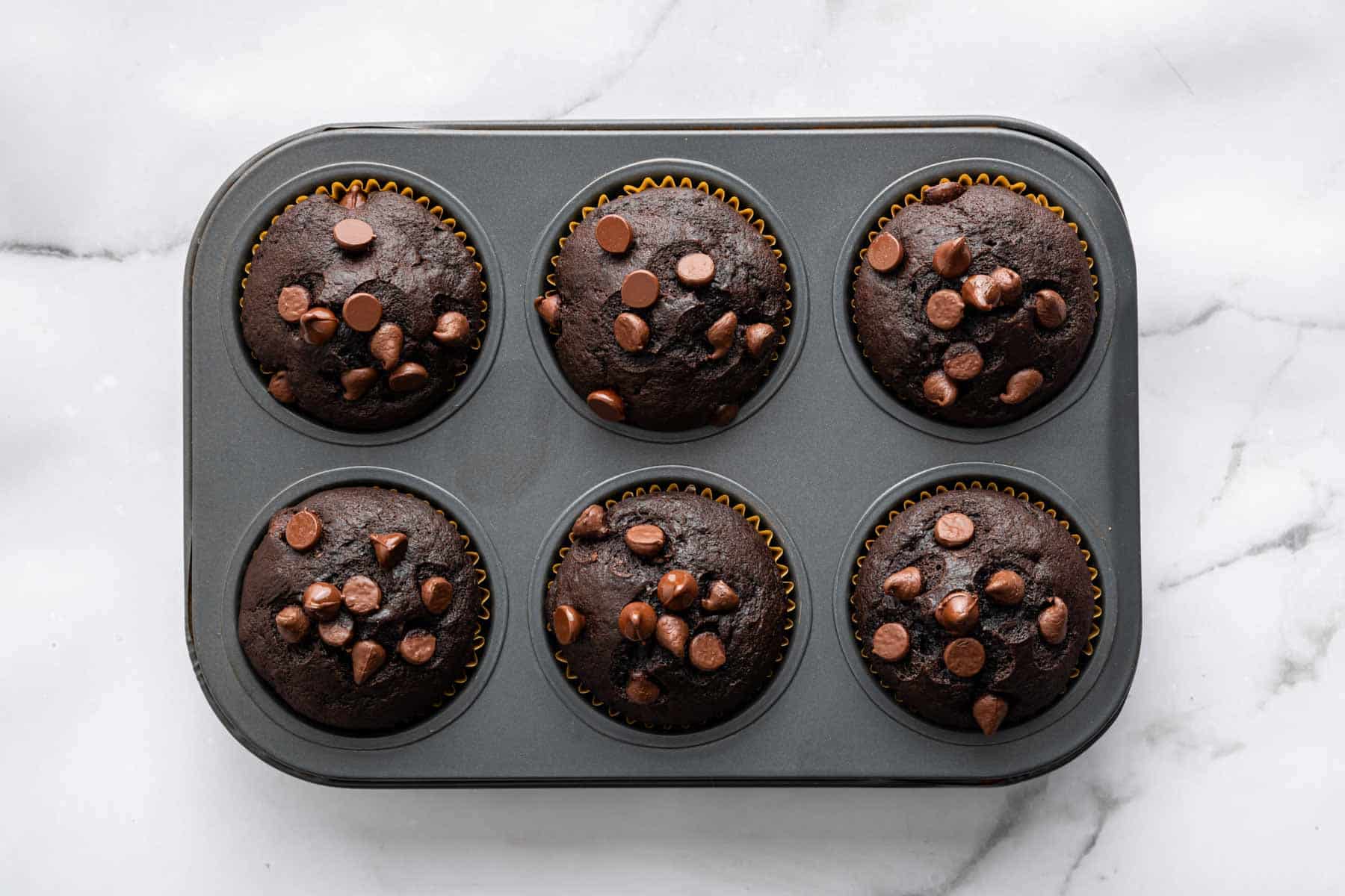 chocolate-banana-muffins Six perfectly domed chocolate banana muffins with chocolate chips in a muffin pan.