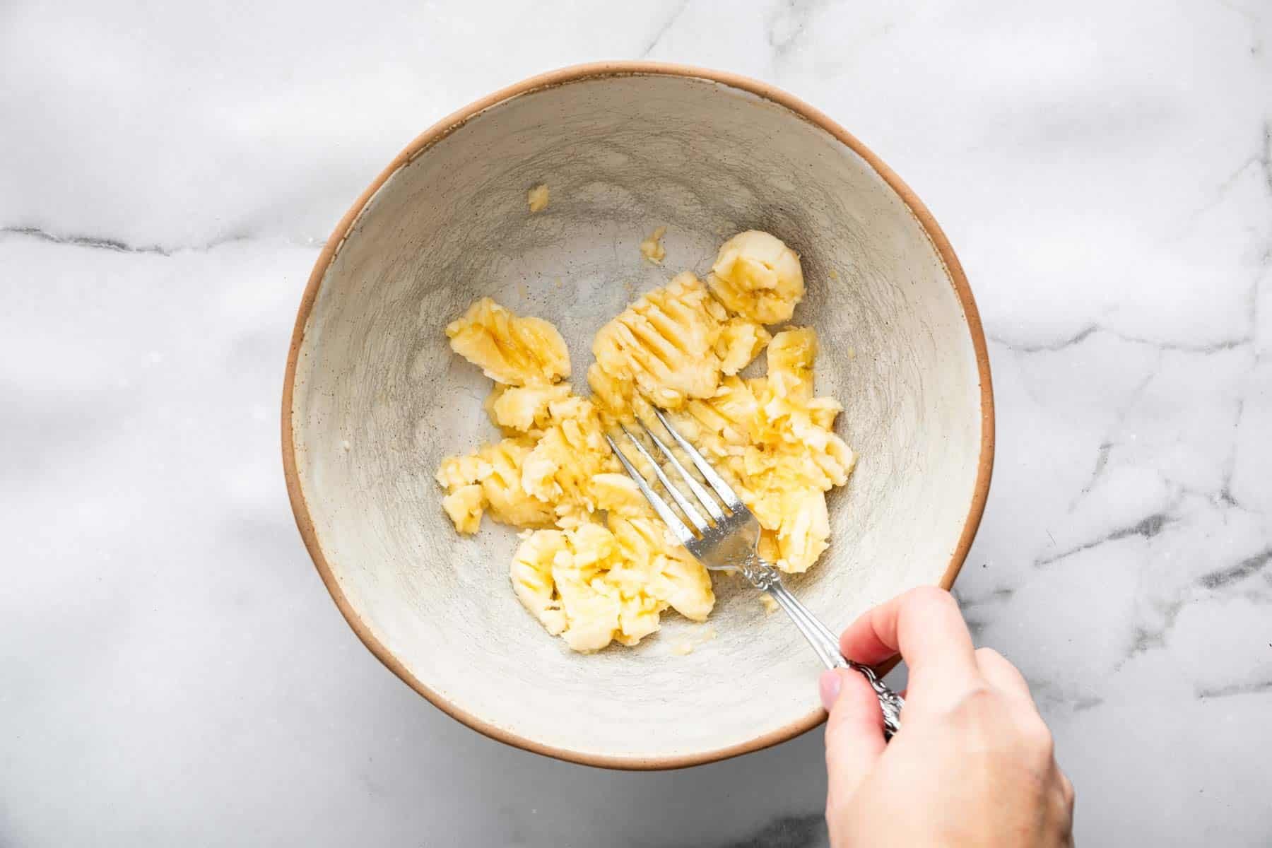 Hand mashing fruit with a fork in a grey bowl.