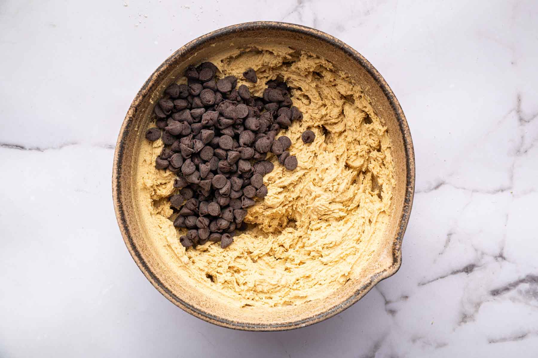 Chocolate chips on top of creamy yellow batter in brown bowl.
