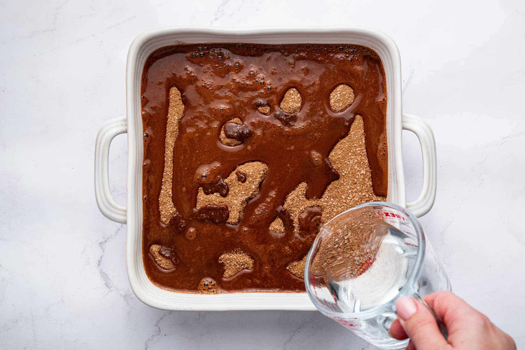 Hand pouring hot water over brown dessert in square pan.