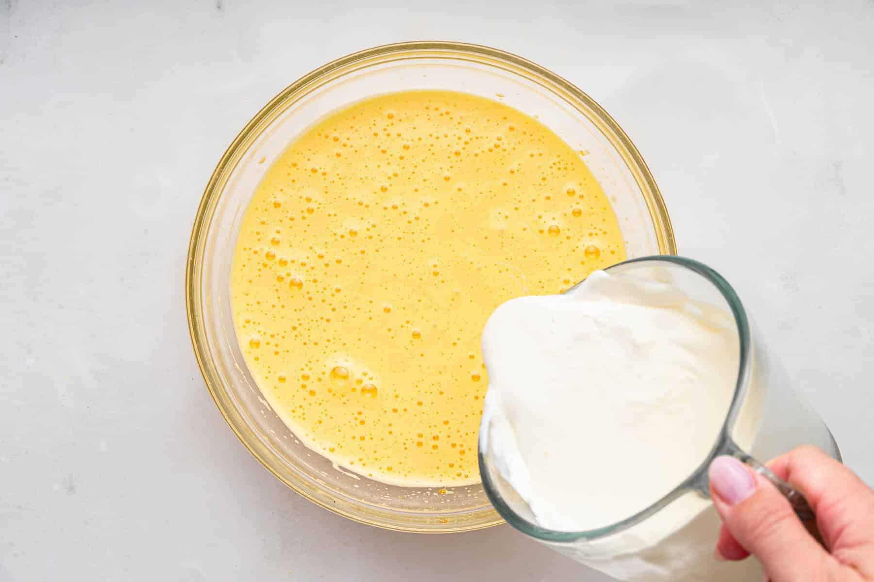 Hand pouring white heavy cream into yellow batter in a bowl.