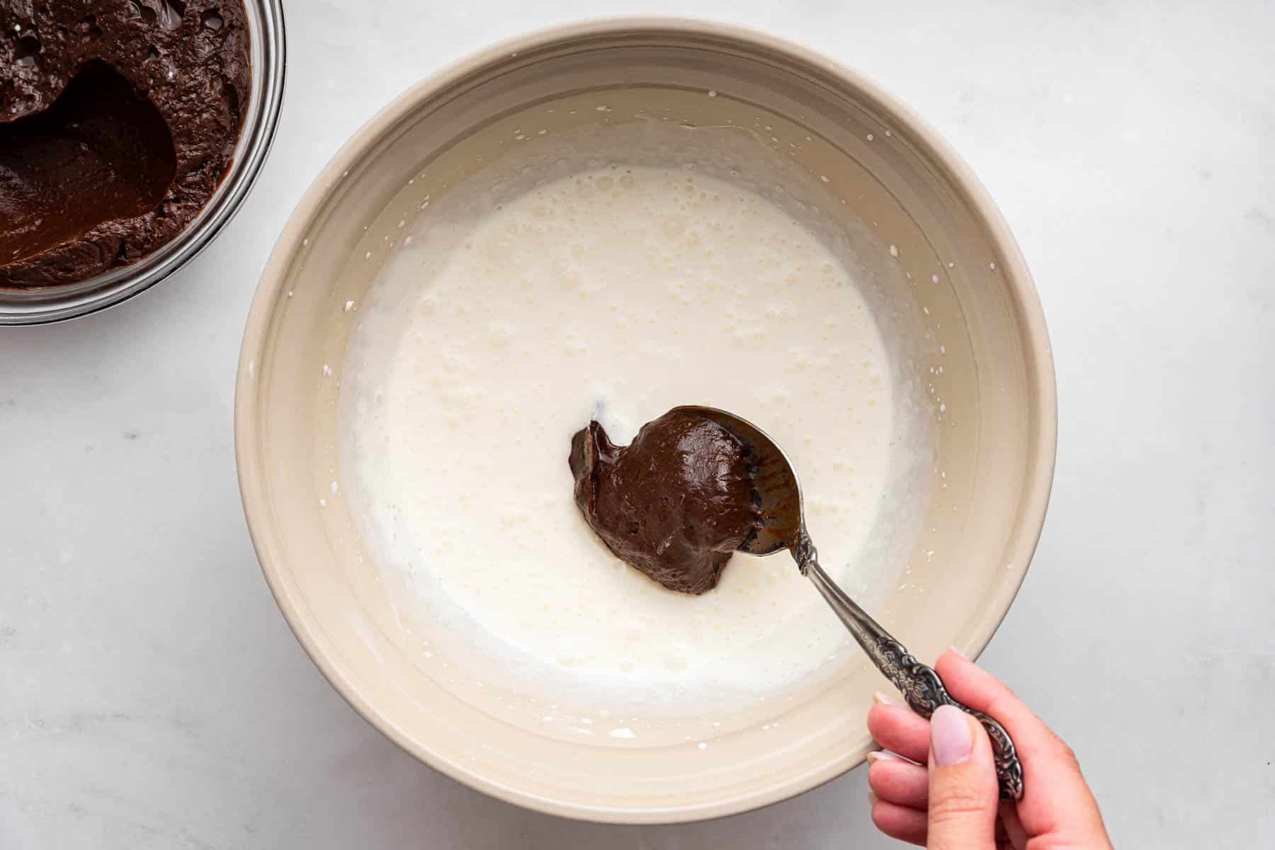 Spooning brown batter into fluffy white whipped cream in a bowl.