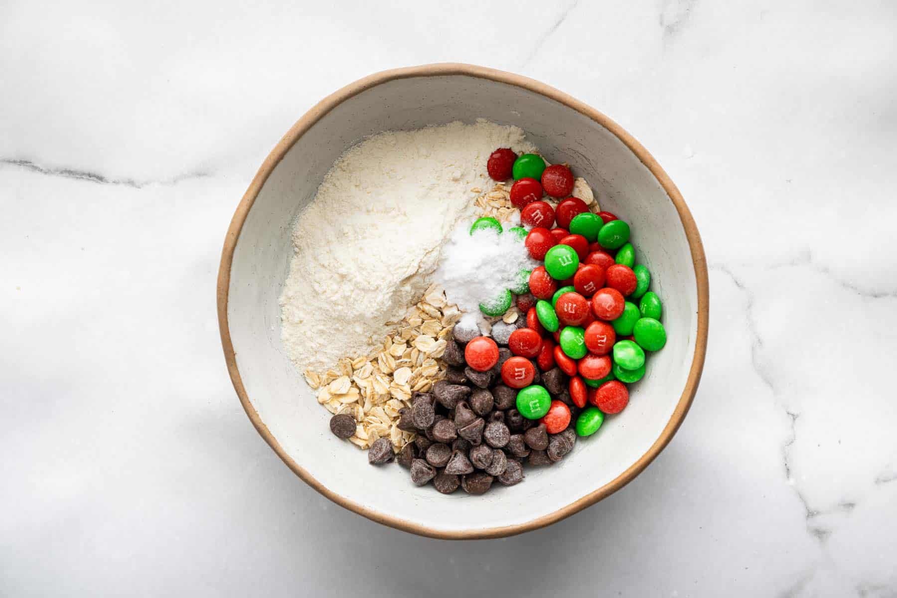 White bowl with flour, oats, chocolate chips, and red and green M&Ms.