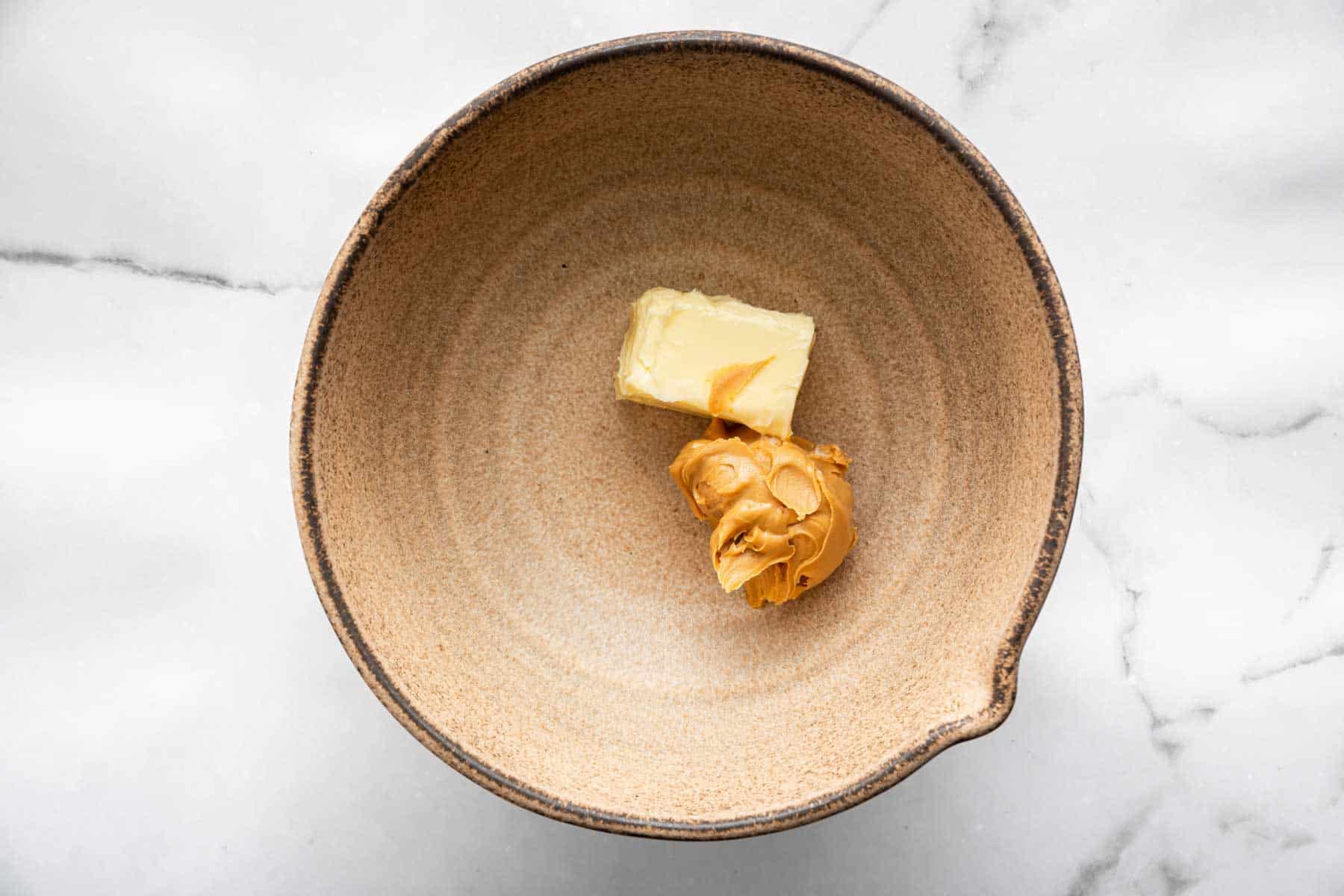 Brown pottery bowl with half a stick of butter and a scoop of peanut butter.