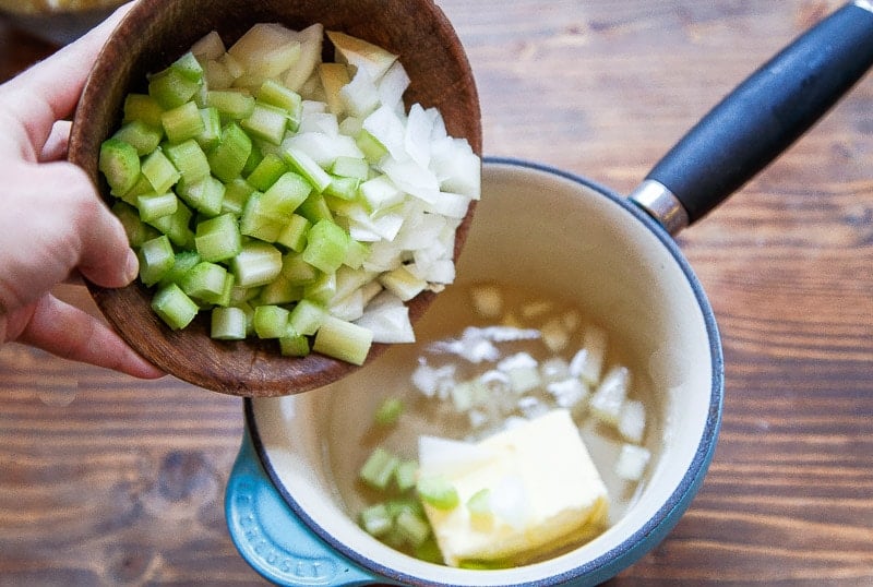 Hand pouring chopped celery and onion into small pan with butter.