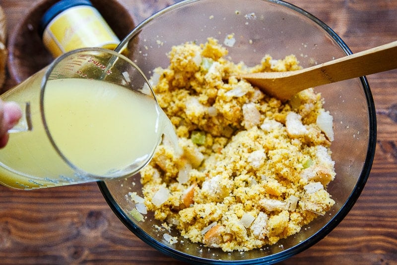 Hand pouring yellow broth into bowl with crumbled bread and spices.