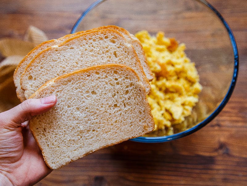 Hand holding sliced bread over a bowl of crumbled yellow bread.
