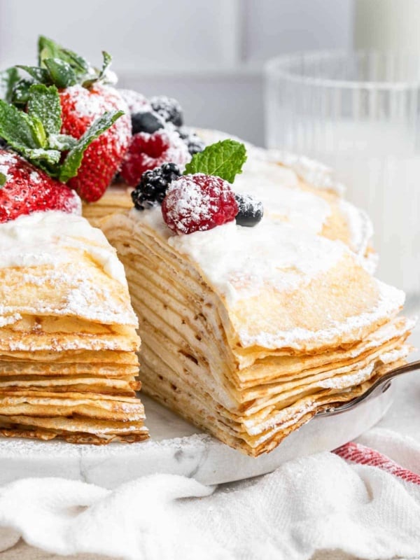 A slice of crepe cake being removed from a whole cake with fresh berries on top.