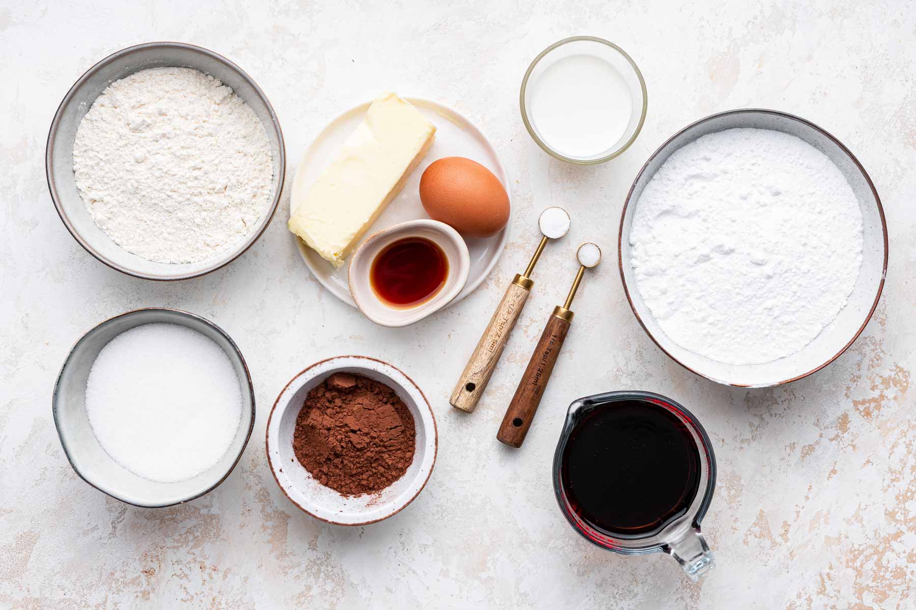 White surface with bowls of sugar, flour, cocoa powder, egg and vanilla.