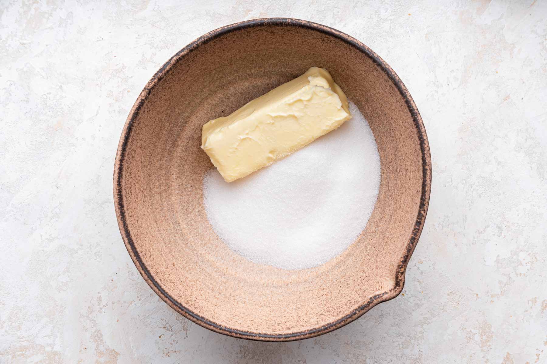 Brown bowl with stick of butter and white granulated sugar in it.