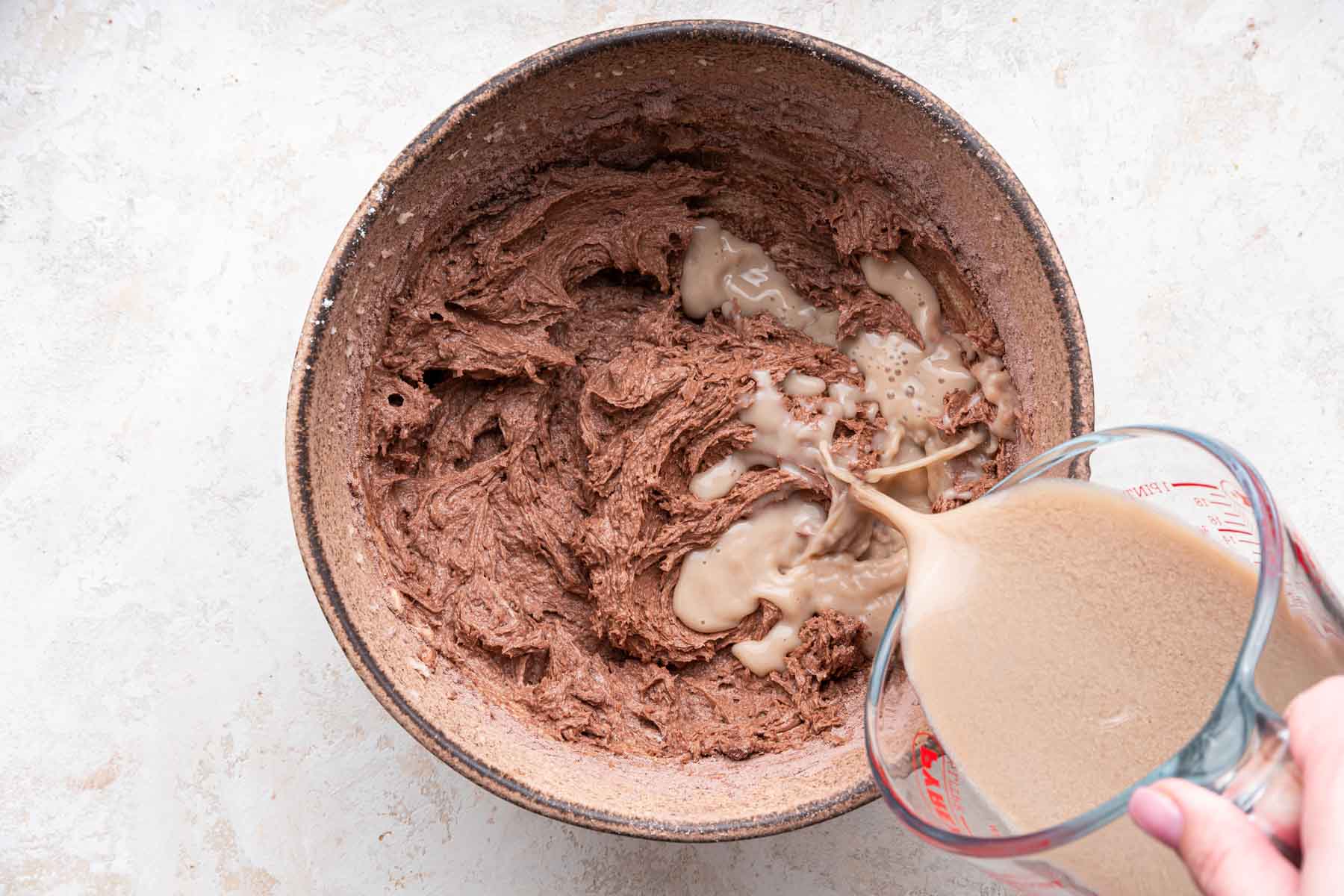 Hand pouring light brown liquid into chocolate cake batter in brown bowl.