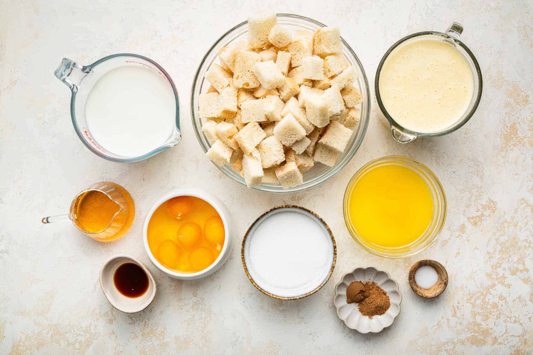 Small bowls with eggs, vanilla, stale bread cubes, milk, and melted butter on kitchen counter.