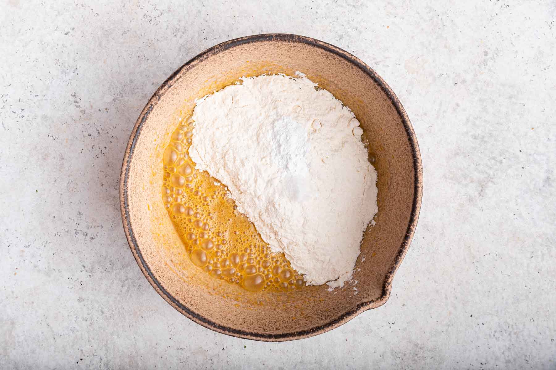 White flour sifted over yellow cake batter in brown bowl.