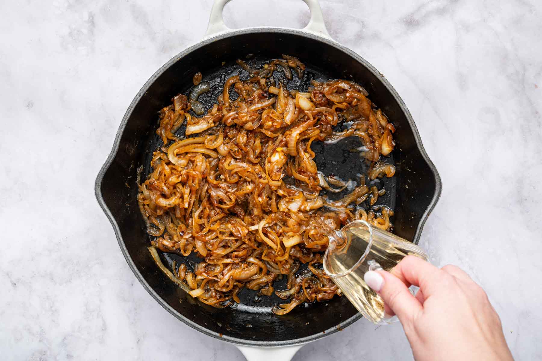 Hand pouring white wine into cast iron skillet with cooked vegetables.
