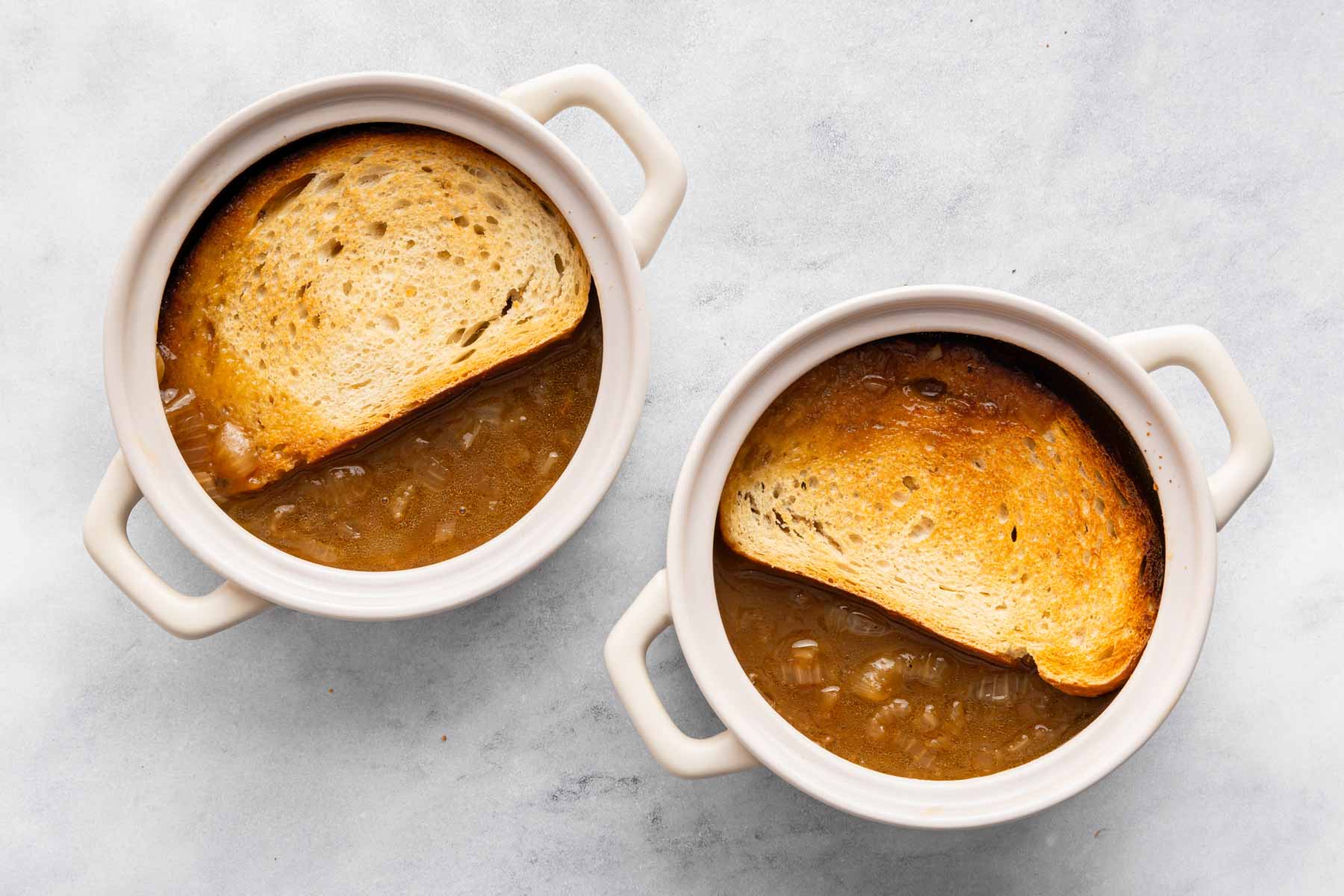 Two bowls of French onion soup with sliced bread on top.