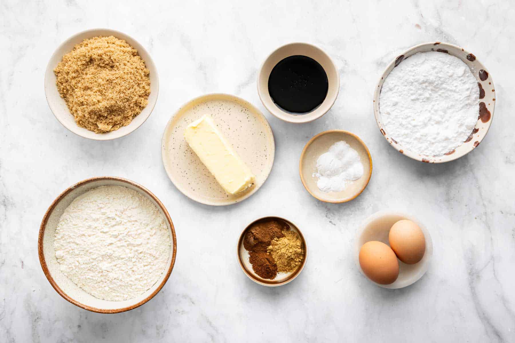 Small bowls of brown sugar, molasses, spices, and eggs on marble counter.