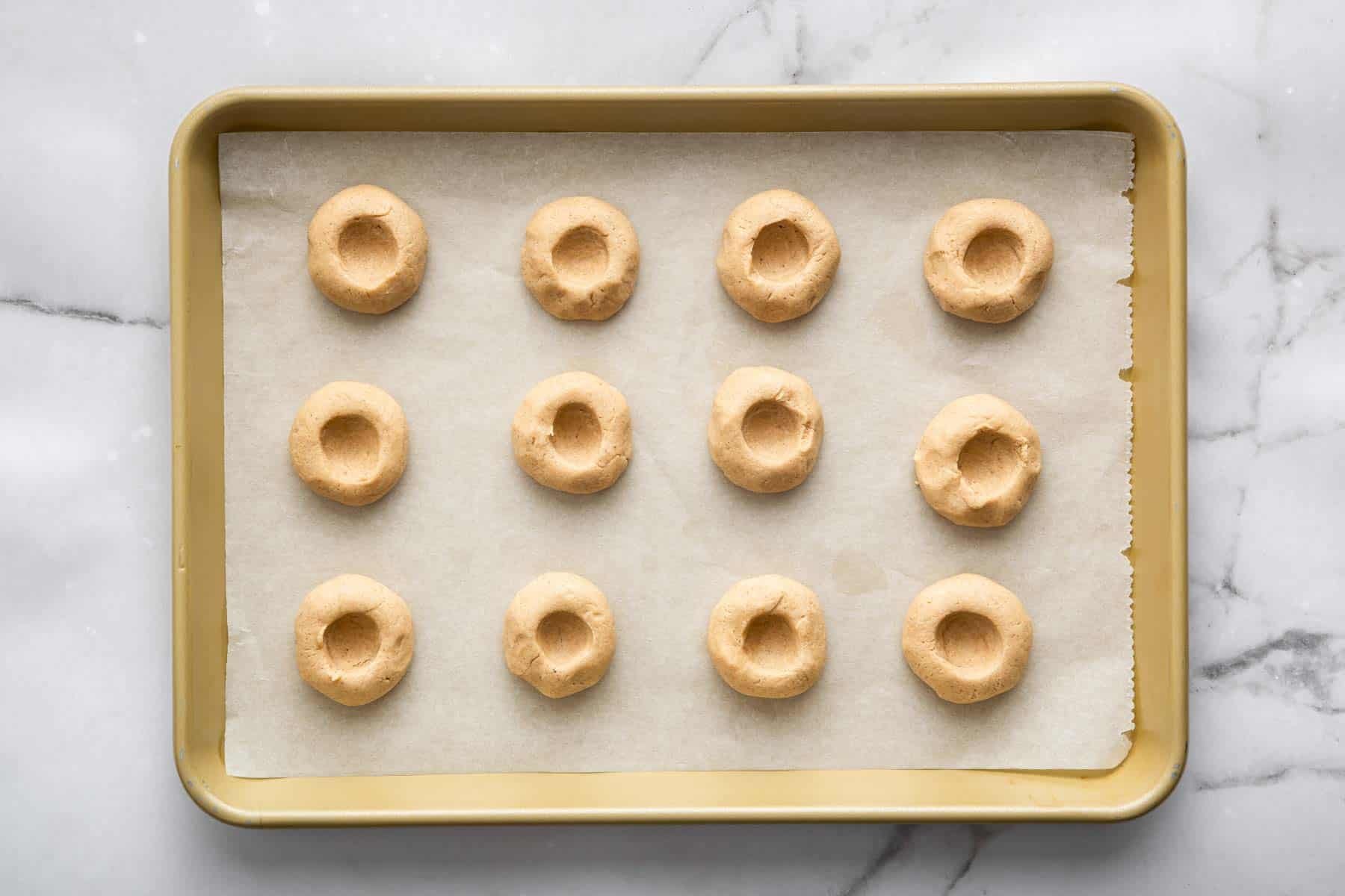 Twelve raw thumbprint cookies on a baking sheet, before baking.
