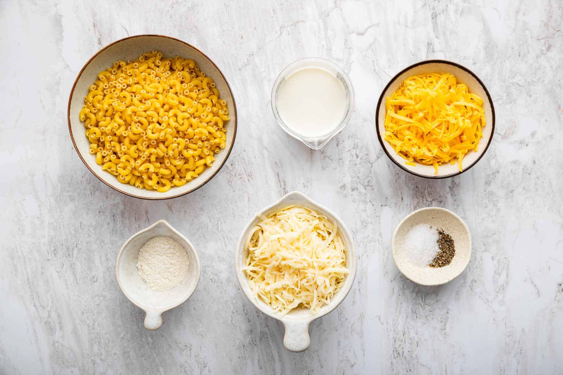 Bowl of noodles, shredded cheddar, milk, and spices on grey counter.