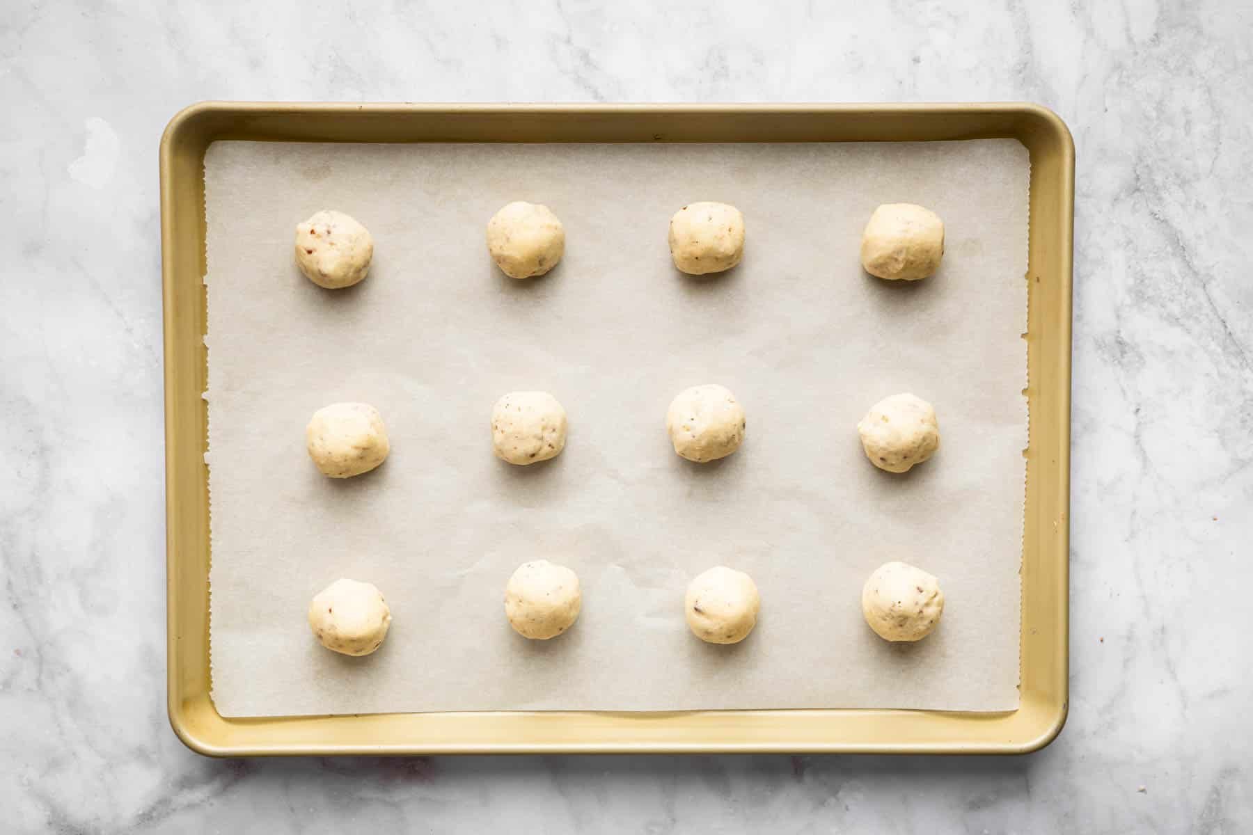 Twelve pale yellow raw cookie dough balls with pecans on baking sheet.