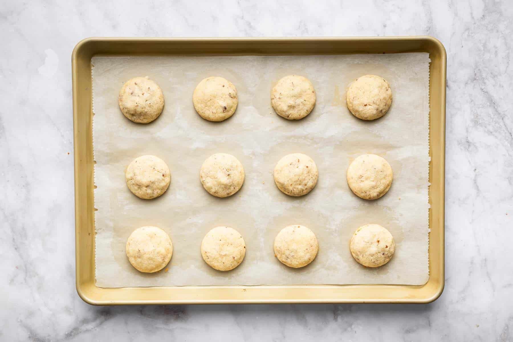 Twelve Mexican wedding cookies on a baking sheet before being rolled in sugar.