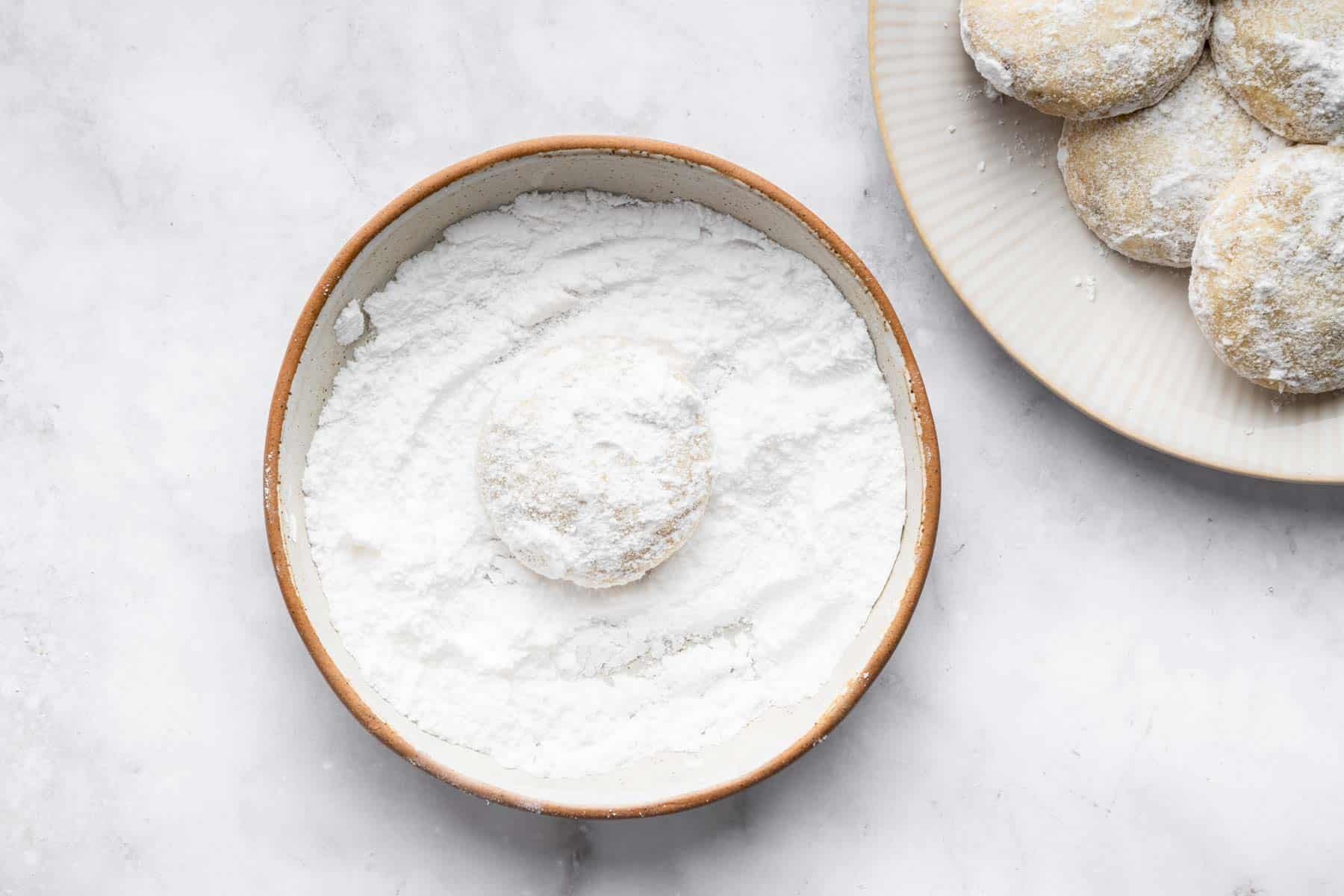 Mexican wedding cookies being rolled in powdered sugar on plate.