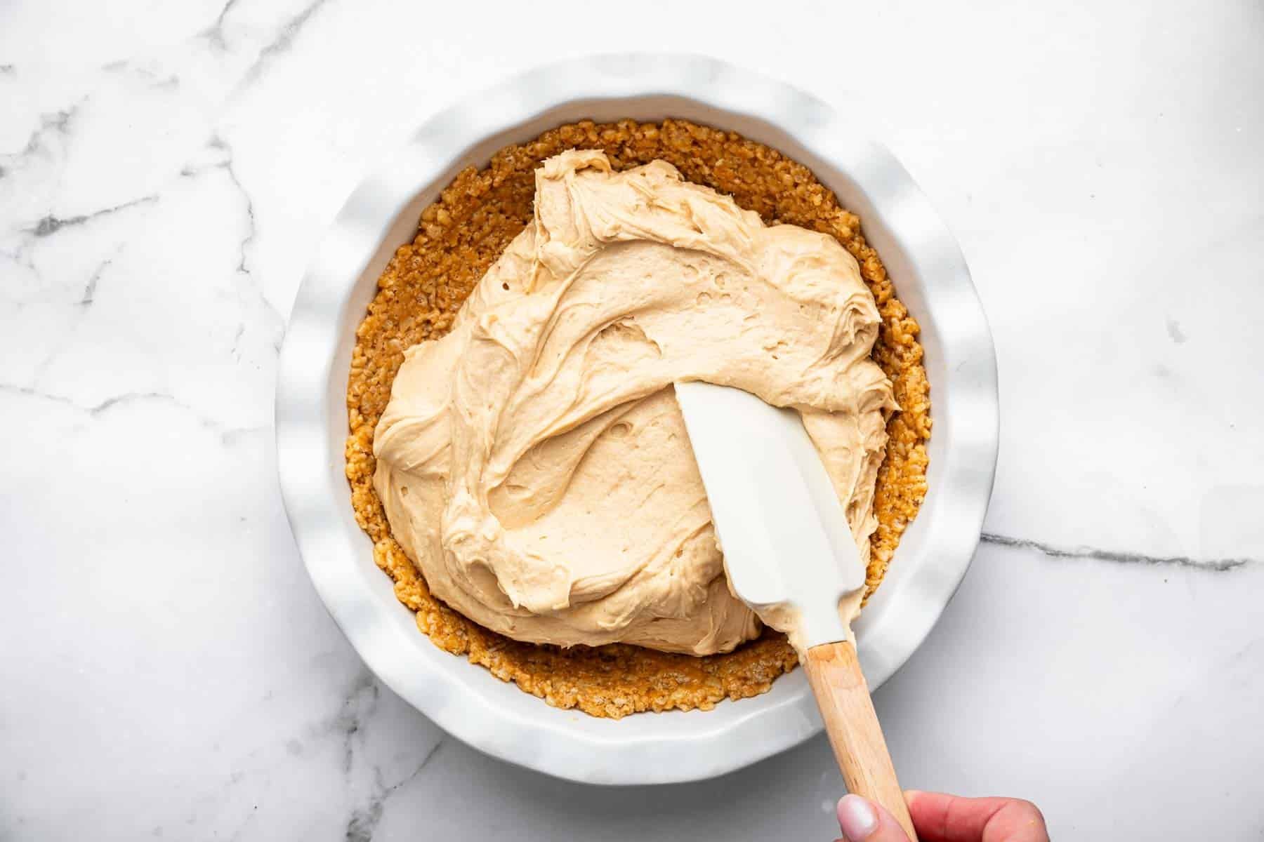 Spatula spreading brown fluff over rice cereal pie crust.