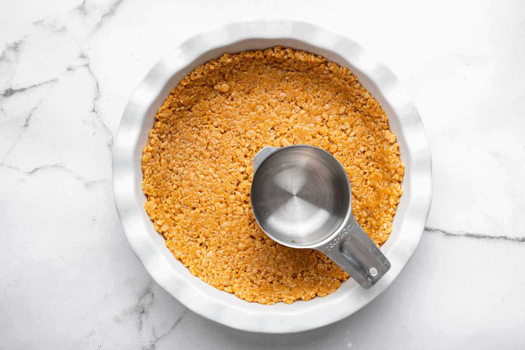 Measuring cup pressing brown rice cereal treats in a pie pan.