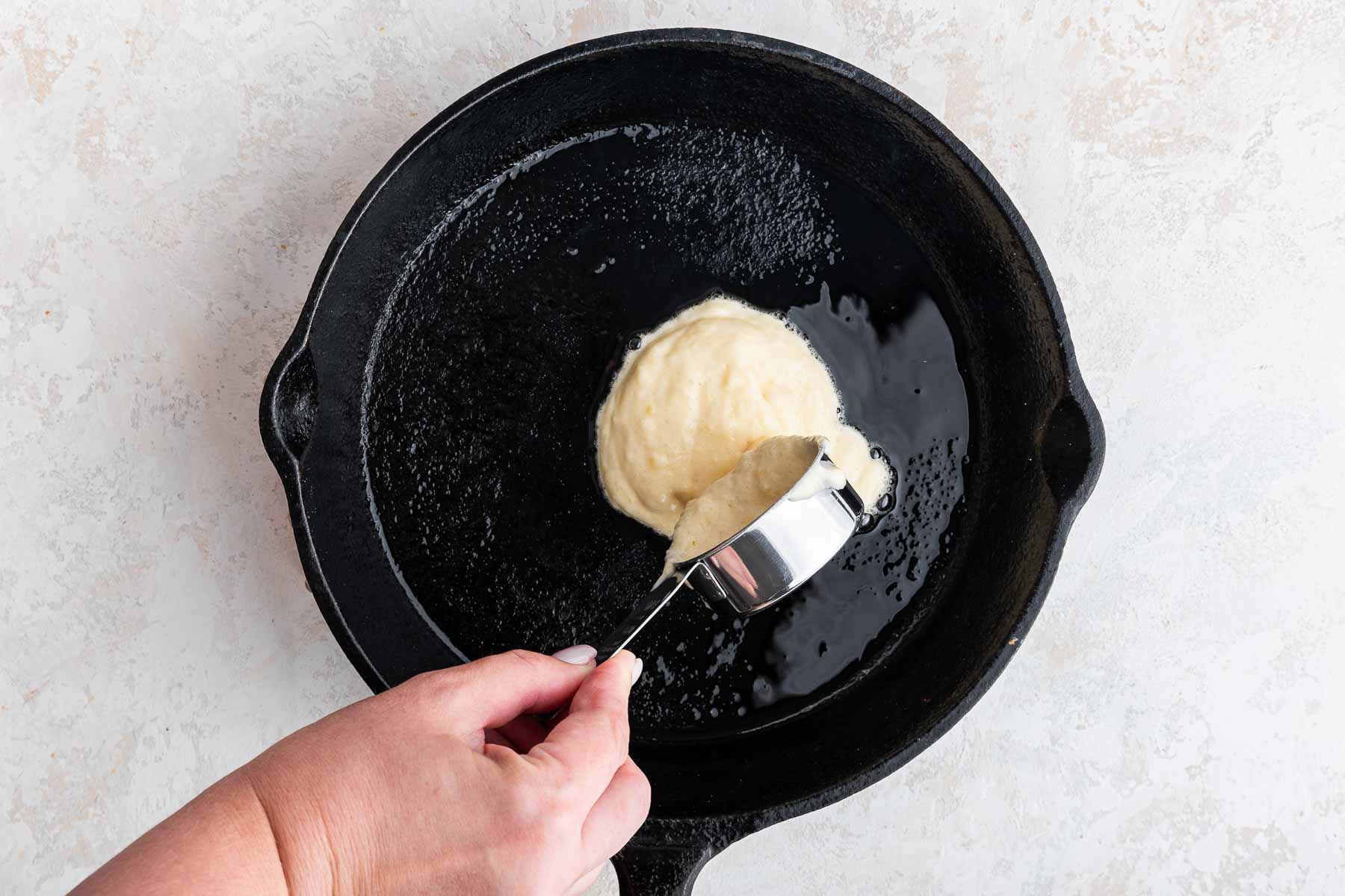 Hand pouring batter into black cast iron skillet from measuring cup.