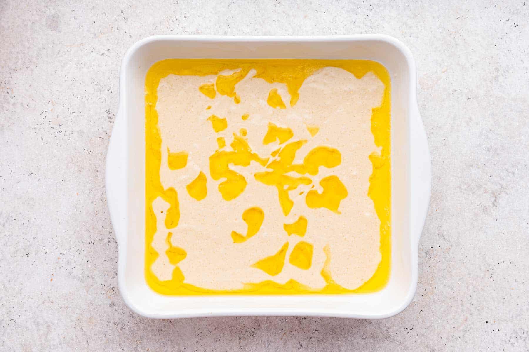 Pancake-like thin batter poured over melted butter in white square baking dish.