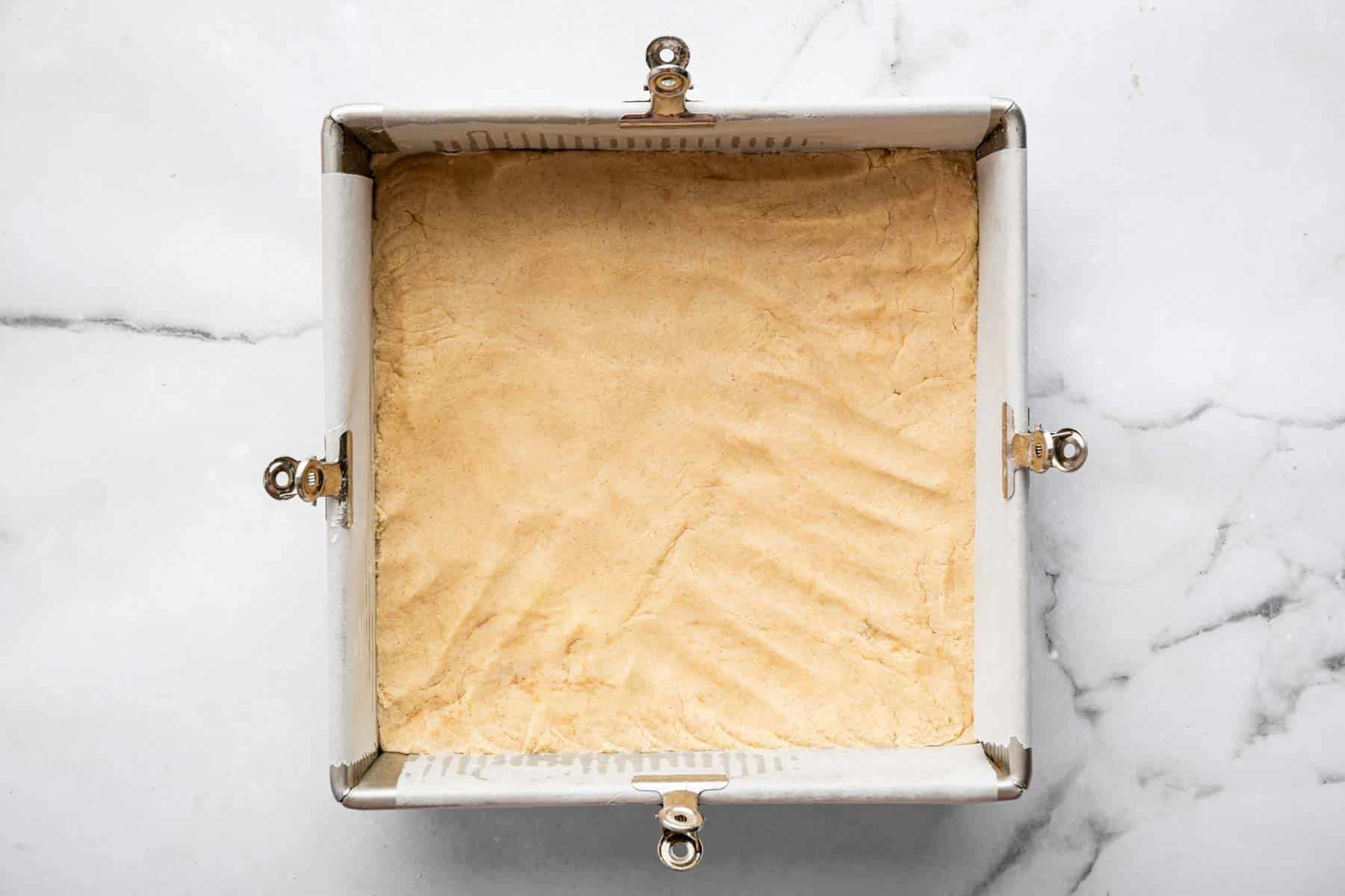 Eight inch square pan with raw shortbread dough pressed into an even layer.