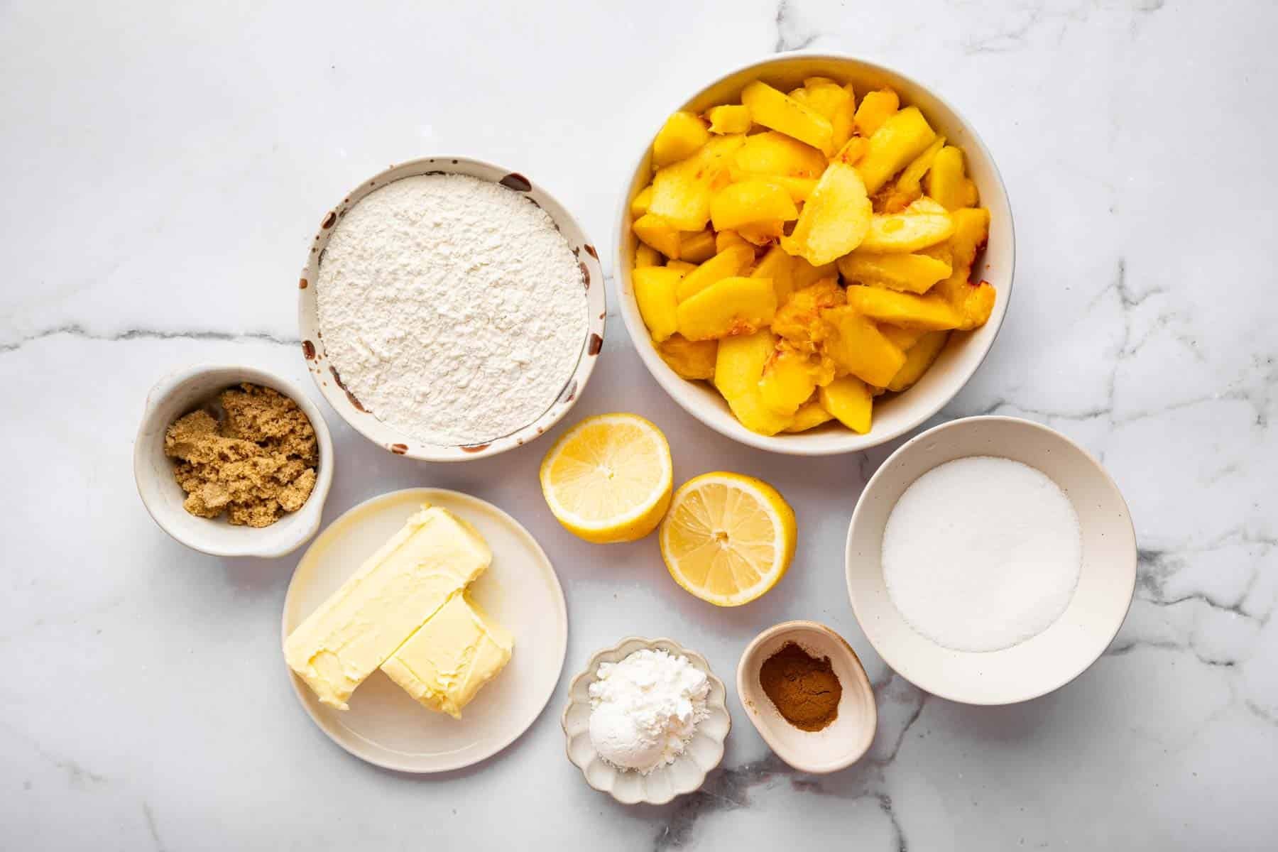 Bowls of sliced fruit, flour, sugar, and butter on marble kitchen counter.
