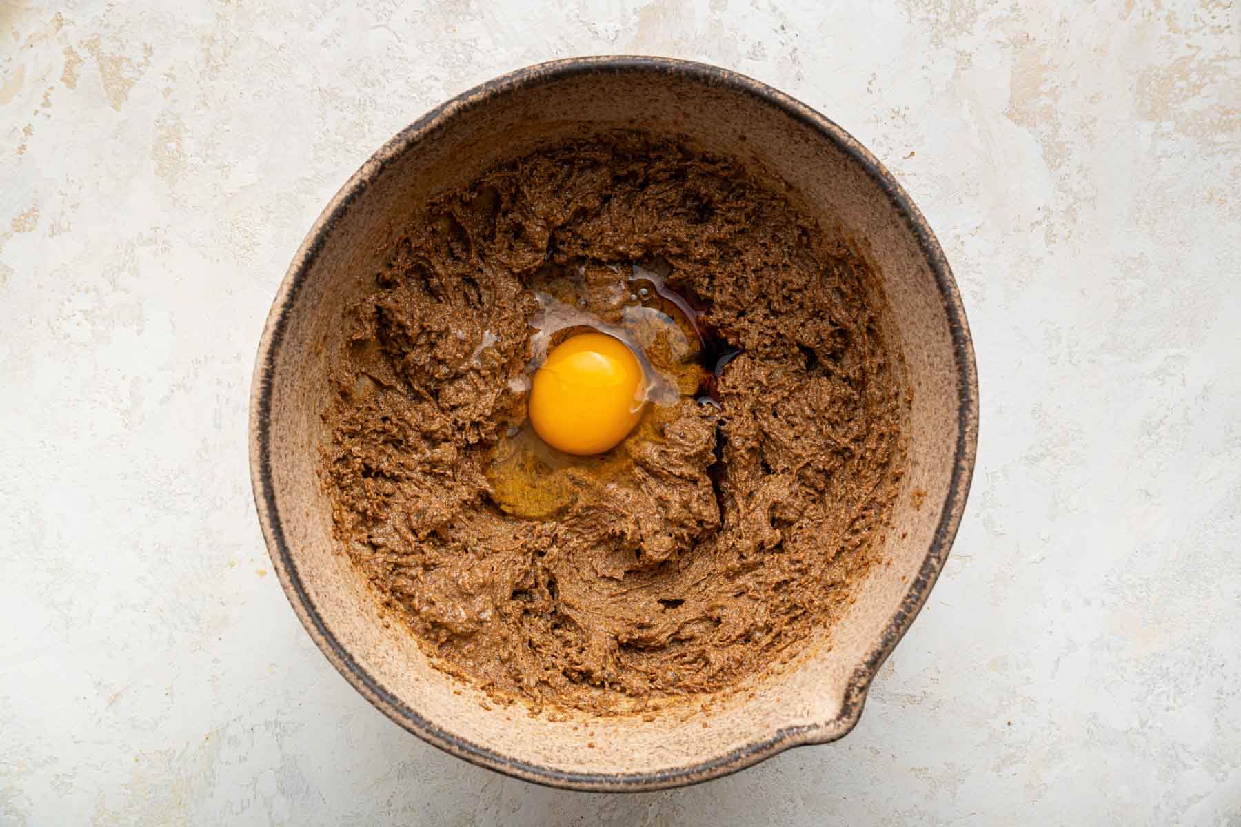 Brown bowl with brown batter and a raw egg and vanilla extract on top.