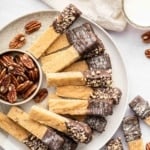 Vertical image of pecan shortbread cookie sticks on a plate with a bowl of pecans.