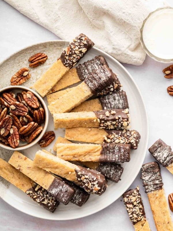 Vertical image of pecan shortbread cookie sticks on a plate with a bowl of pecans.