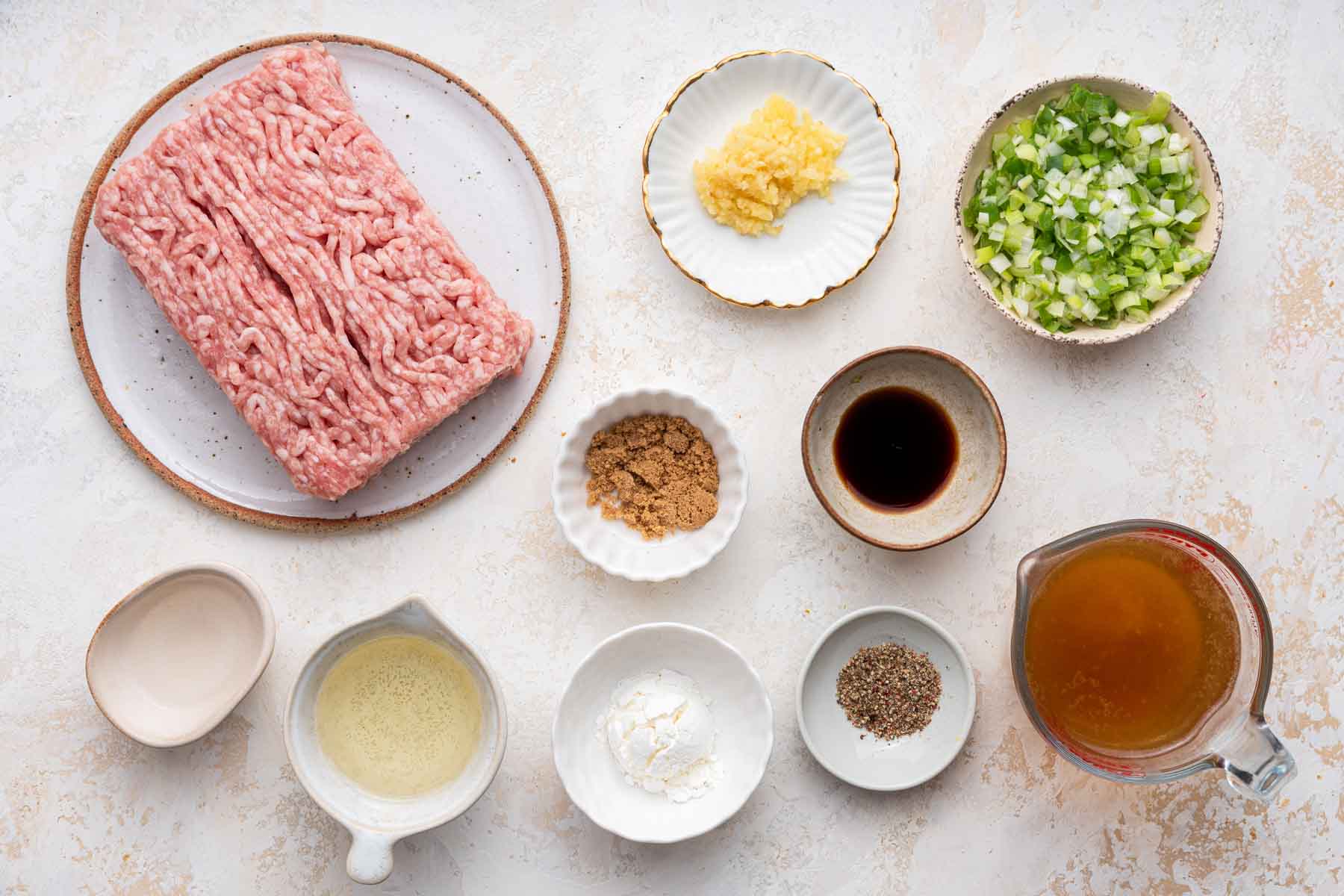 Ground pork, chopped garlic, soy, and scallions in small bowls on white counter.
