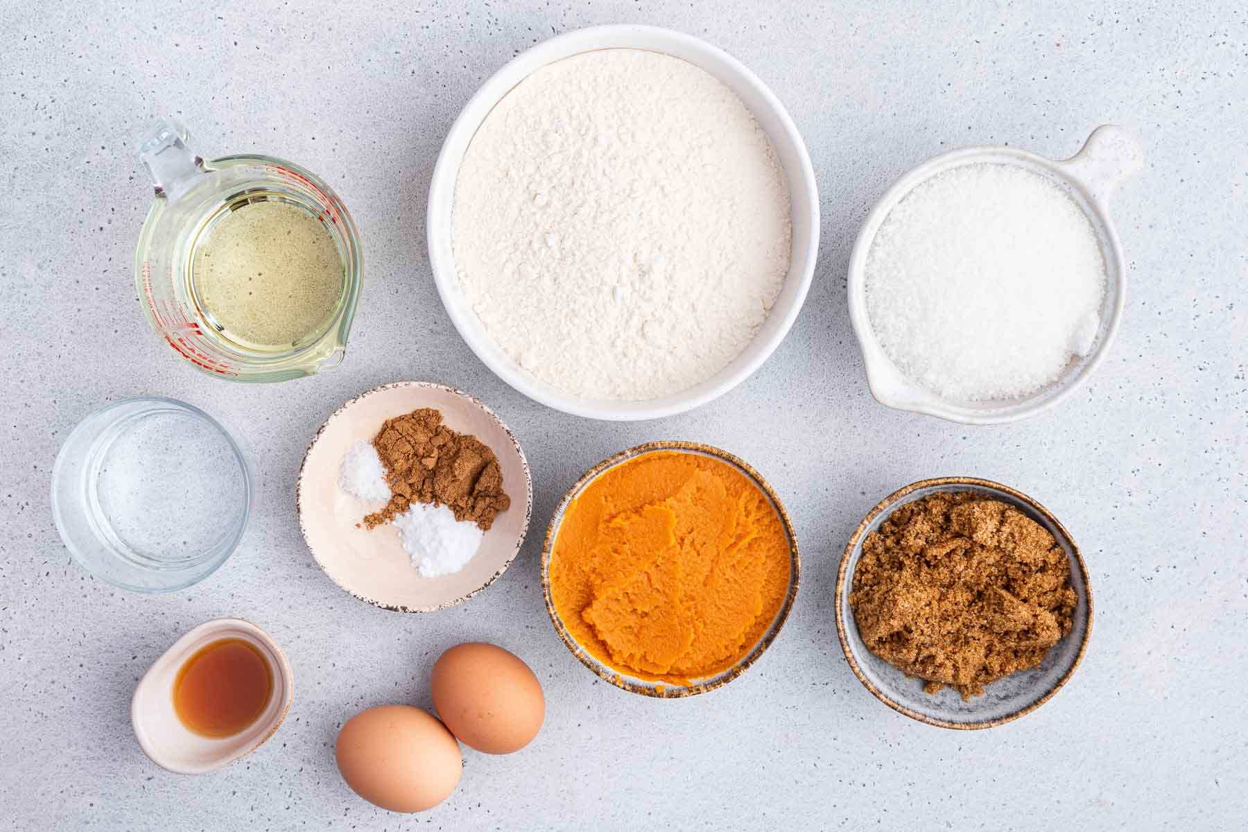 Small bowls of flour, sugar, canned orange squash, brown sugar and eggs on kitchen surface.